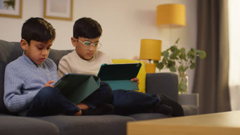 Two-Young-Boys-Sitting-On-Sofa-At-Home-Playing-Games-Or-Streaming-Onto-Digital-Tablets-14